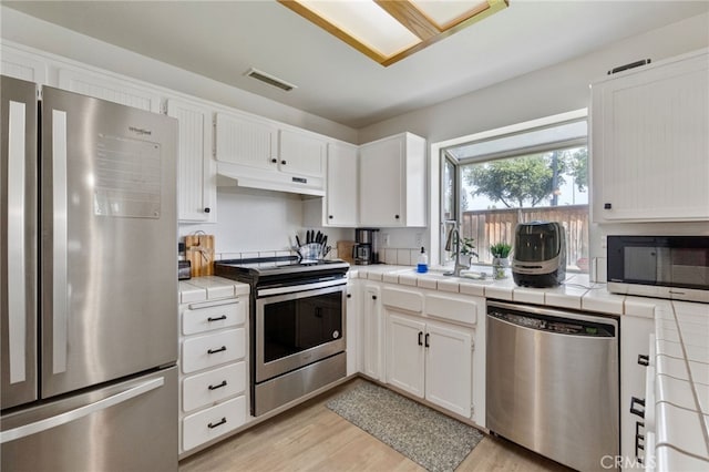 kitchen featuring appliances with stainless steel finishes, under cabinet range hood, tile countertops, light wood-type flooring, and white cabinets