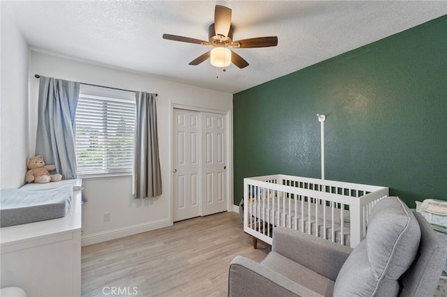 bedroom featuring light wood finished floors, ceiling fan, baseboards, a crib, and a textured ceiling