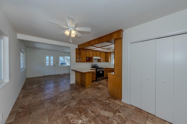 kitchen with stainless steel gas range oven, under cabinet range hood, brown cabinetry, stone finish flooring, and light countertops