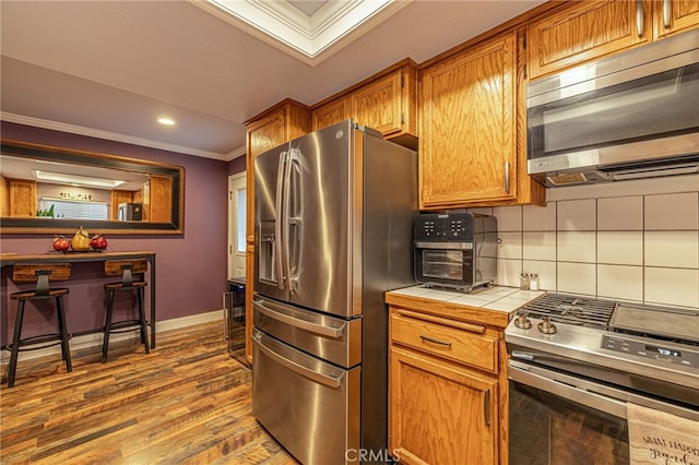 43156 41st St W, Lancaster, CA 93536 - kitchen featuring appliances with stainless steel finishes, tile countertops, ornamental molding, wood finished floors, and brown cabinets