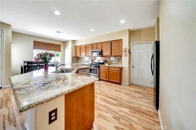 kitchen with appliances with stainless steel finishes, a sink, light wood-type flooring, brown cabinetry, and an island with sink