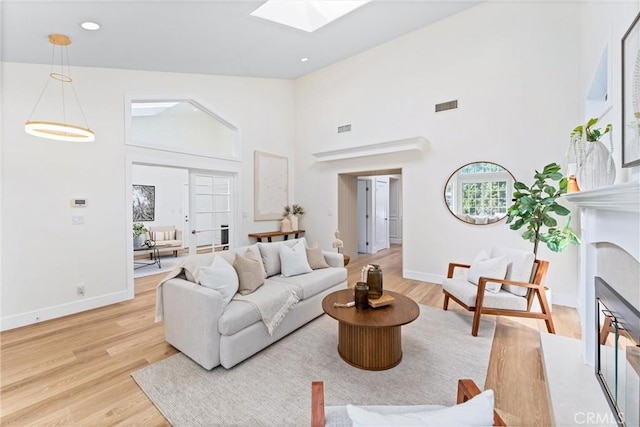 living area featuring light wood-style floors, a skylight, high vaulted ceiling, baseboards, and a fireplace with flush hearth