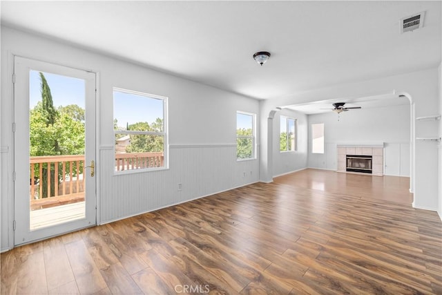 unfurnished living room with a ceiling fan, wood finished floors, a tiled fireplace, and wainscoting