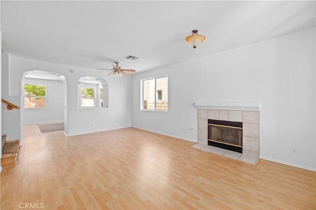 unfurnished living room featuring light wood finished floors, a tile fireplace, a ceiling fan, arched walkways, and baseboards