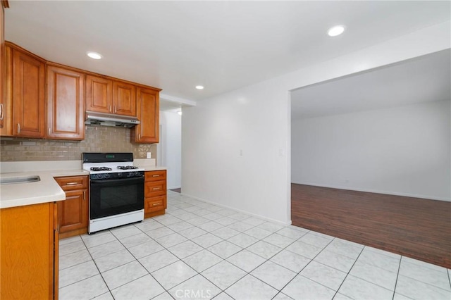 kitchen with white range with gas cooktop, under cabinet range hood, light countertops, backsplash, and brown cabinetry