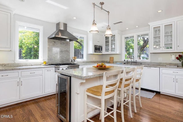 kitchen featuring wine cooler, white dishwasher, wall chimney range hood, a sink, and white cabinetry