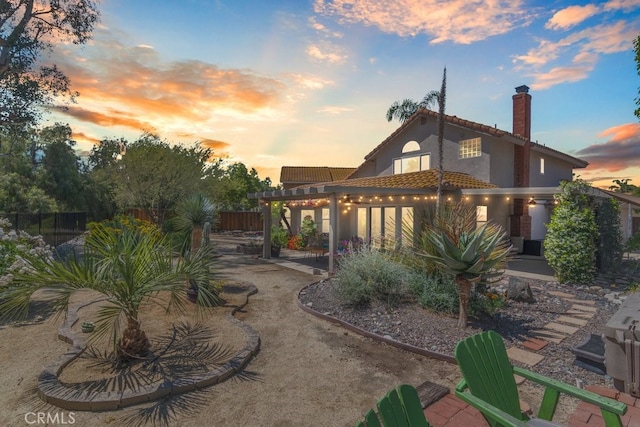 back of property at dusk with a tile roof, a chimney, fence, stucco siding, and a pergola