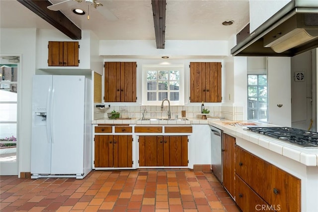 kitchen with stainless steel appliances, beam ceiling, wall chimney range hood, tile counters, and brown cabinetry
