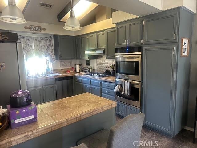 11420 Lasselle St, Moreno Valley, CA 92557 - kitchen featuring tile counters, appliances with stainless steel finishes, under cabinet range hood, backsplash, and vaulted ceiling