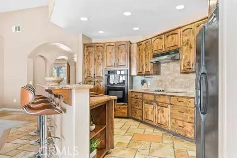 62398 Cope Ln, Joshua Tree, CA 92252 - kitchen with under cabinet range hood, black appliances, backsplash, brown cabinetry, and recessed lighting