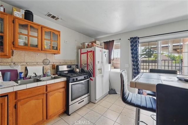 14276 Aztec St, Sylmar, CA 91342 - kitchen with stainless steel range with gas stovetop, tile countertops, light tile patterned floors, brown cabinetry, and white refrigerator with ice dispenser