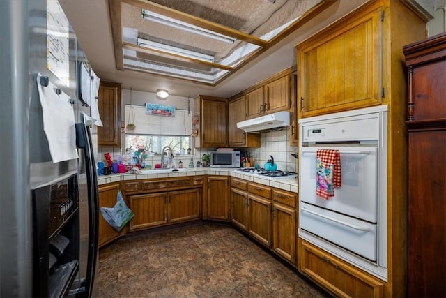 56455 Carlyle Dr, Yucca Valley, CA 92284 - kitchen with white appliances, a warming drawer, under cabinet range hood, tile counters, and brown cabinets