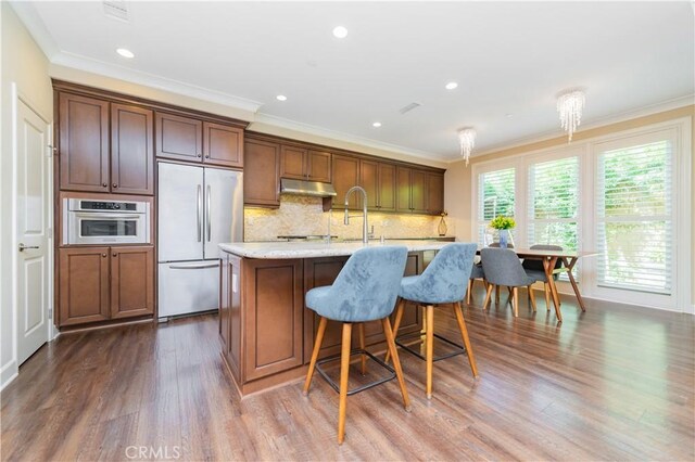 43 Lupari, Irvine, CA 92618 - kitchen featuring appliances with stainless steel finishes, under cabinet range hood, dark wood-type flooring, a kitchen island with sink, and decorative backsplash