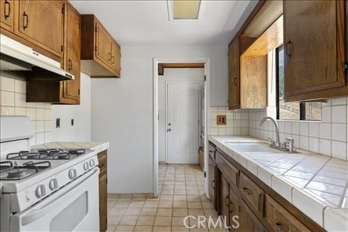 28921 Bear Valley Rd, Tehachapi, CA 93561 - kitchen featuring white gas range oven, tile counters, a sink, under cabinet range hood, and tasteful backsplash