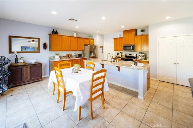 31786 Sorrel Run Ct, Menifee, CA 92584 - kitchen with appliances with stainless steel finishes, brown cabinets, a kitchen island with sink, light stone counters, and light tile patterned floors