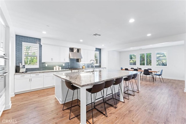 17150 Vita Way, Riverside, CA 92503 - kitchen featuring wall chimney range hood, a breakfast bar, a kitchen island with sink, light stone counters, and white cabinetry
