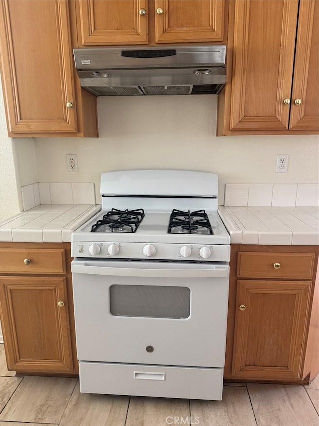 14543 Handsdale St, Adelanto, CA 92301 - kitchen featuring white gas range oven, under cabinet range hood, tile counters, and brown cabinets