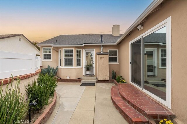 6003 Eastbrook Ave, Lakewood, CA 90713 - exterior entry at dusk featuring stucco siding, a patio area, a chimney, and a shingled roof