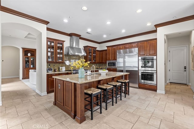 19103 Green Oaks Rd, Yorba Linda, CA 92886 - kitchen featuring wall chimney exhaust hood, built in appliances, backsplash, a center island with sink, and a breakfast bar area