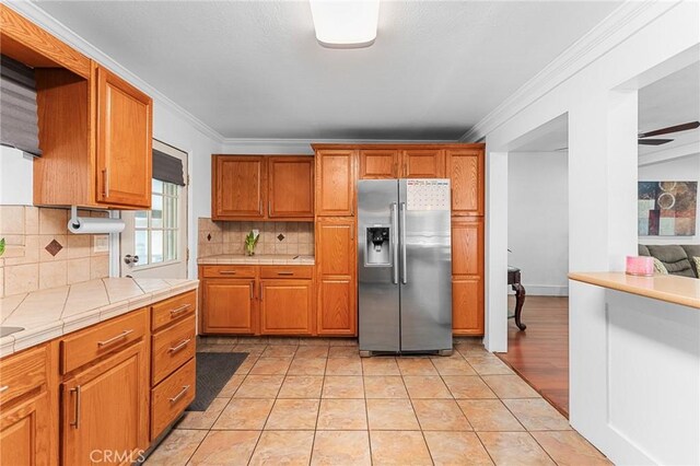 1040 W 3rd St, Pomona, CA 91766 - kitchen with stainless steel fridge, tile countertops, decorative backsplash, brown cabinetry, and ornamental molding