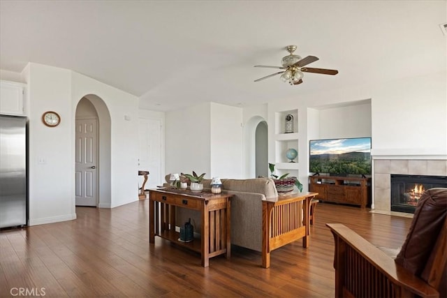 1467 Hunter Moon Way, Beaumont, CA 92223 - living room featuring arched walkways, a ceiling fan, a tiled fireplace, built in shelves, and dark wood-style floors