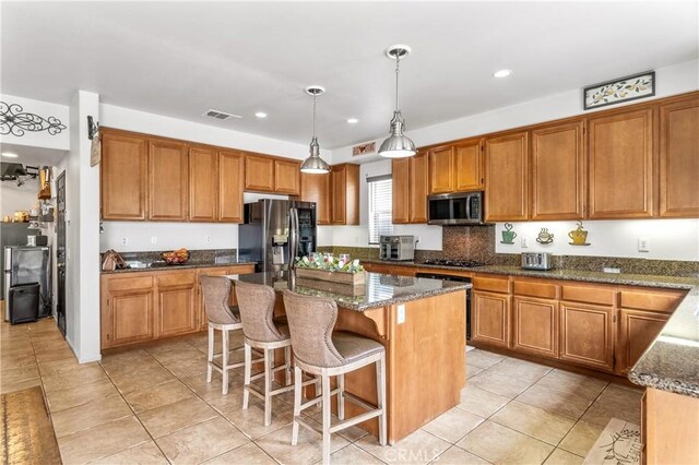 3568 Rock Butte Pl, Perris, CA 92570 - kitchen featuring appliances with stainless steel finishes, dark stone countertops, a kitchen island, light tile patterned flooring, and brown cabinetry