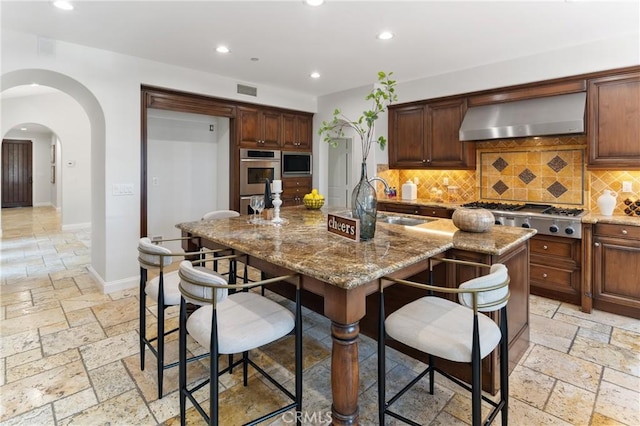 15 Basilica Pl, Ladera Ranch, CA 92694 - kitchen featuring wall chimney range hood, stainless steel gas stovetop, stone tile floors, recessed lighting, and arched walkways