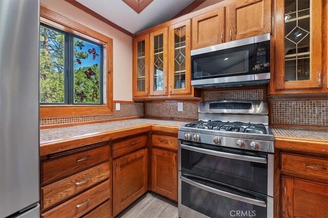 26745 Hopkins Rd, Idyllwild, CA 92549 - kitchen featuring appliances with stainless steel finishes, backsplash, brown cabinetry, glass insert cabinets, and light wood finished floors