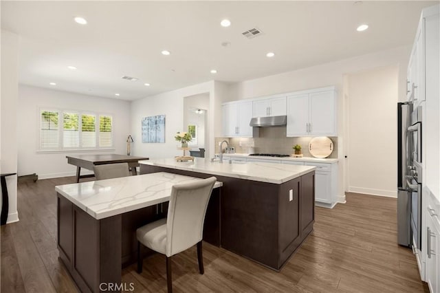 11638 Oakton Way, Corona, CA 92883 - kitchen with under cabinet range hood, a kitchen island with sink, dark wood-style flooring, tasteful backsplash, and white cabinetry