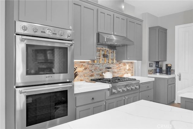 7 Olympic Way, Coto De Caza, CA 92679 - kitchen with appliances with stainless steel finishes, gray cabinetry, under cabinet range hood, light stone countertops, and decorative backsplash