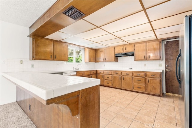 42159 Mayberry Ave, Hemet, CA 92544 - kitchen featuring under cabinet range hood, a peninsula, freestanding refrigerator, brown cabinetry, and decorative backsplash