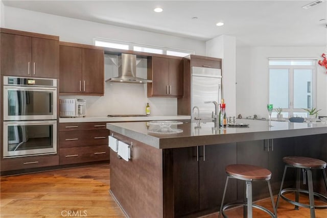 5823 Seahorse Ct, Playa Vista, CA 90094 - kitchen featuring appliances with stainless steel finishes, wall chimney exhaust hood, a breakfast bar, light wood-style floors, and a center island with sink