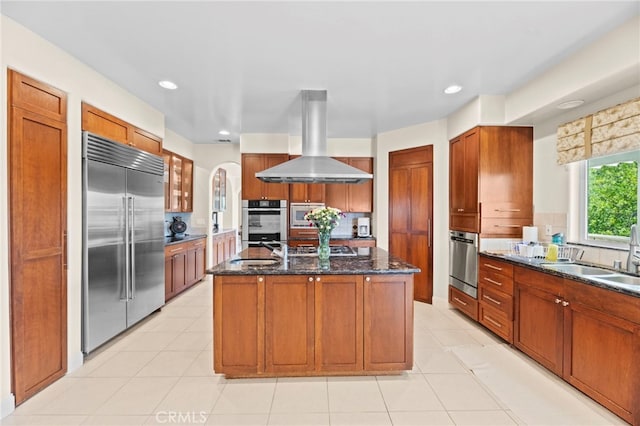 43845 Calle De Velardo, Temecula, CA 92592 - kitchen with appliances with stainless steel finishes, wall chimney exhaust hood, brown cabinetry, dark stone countertops, and a center island with sink