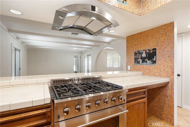45740 Verba Santa Dr, Palm Desert, CA 92260 - kitchen featuring stainless steel stove, tile counters, island exhaust hood, light tile patterned flooring, and beamed ceiling