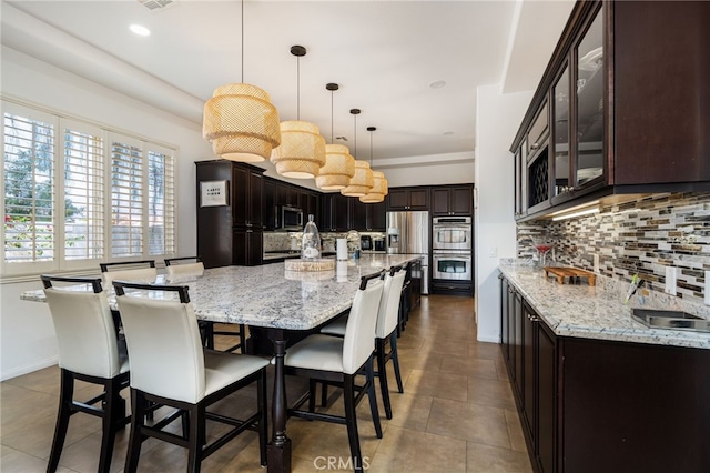43875 Spiaggia Pl, Indio, CA 92203 - kitchen featuring stainless steel appliances, backsplash, a large island with sink, dark brown cabinets, and light stone counters