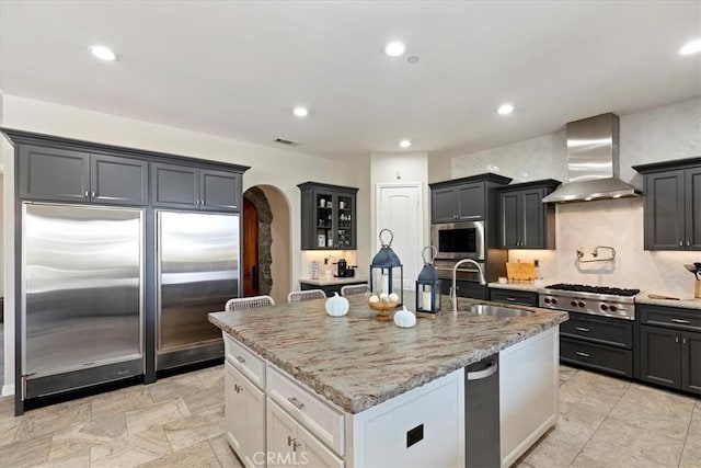 17180 Mockingbird Canyon Rd, Riverside, CA 92504 - kitchen featuring stainless steel appliances, wall chimney exhaust hood, arched walkways, light stone countertops, and a kitchen island with sink