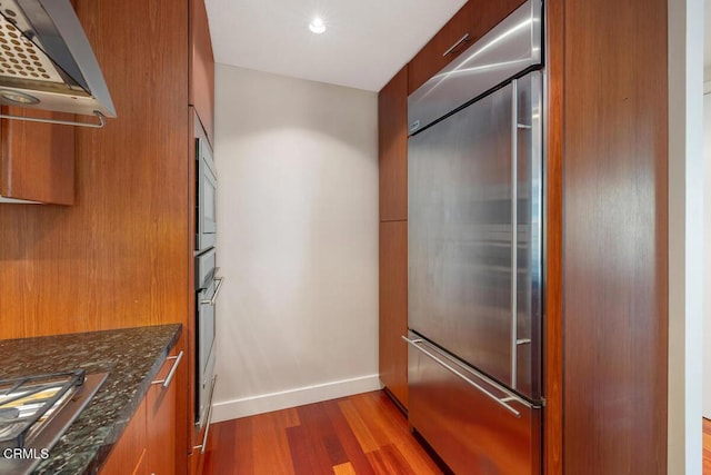 1205 Pacific Hwy Unit 1005, San Diego, CA 92101 - kitchen featuring built in appliances, wall chimney range hood, brown cabinetry, dark wood-style flooring, and dark stone counters