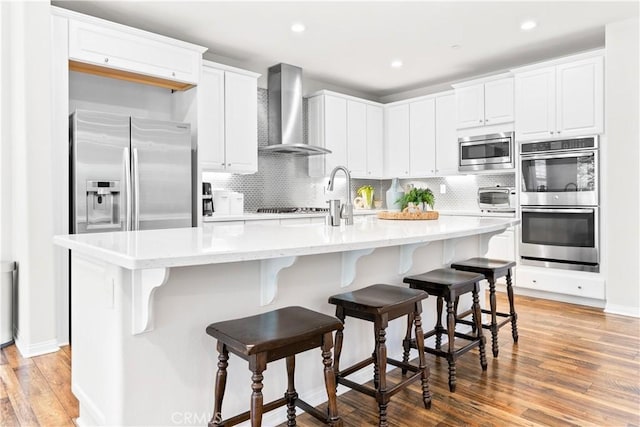 27 Cache St, Rancho Mission Viejo, CA 92694 - kitchen featuring wall chimney range hood, stainless steel appliances, light wood-style floors, decorative backsplash, and an island with sink
