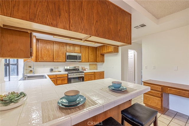 44118 Ruthron Ave, Lancaster, CA 93536 - kitchen with appliances with stainless steel finishes, brown cabinetry, light tile patterned floors, tile counters, and a peninsula