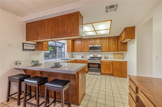 44118 Ruthron Ave, Lancaster, CA 93536 - kitchen featuring stainless steel appliances, brown cabinetry, a peninsula, light tile patterned floors, and a breakfast bar area