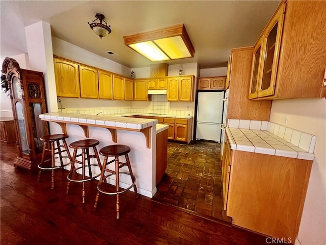 21509 Stage Dr, Tehachapi, CA 93561 - kitchen featuring freestanding refrigerator, a peninsula, tile countertops, a breakfast bar, and under cabinet range hood