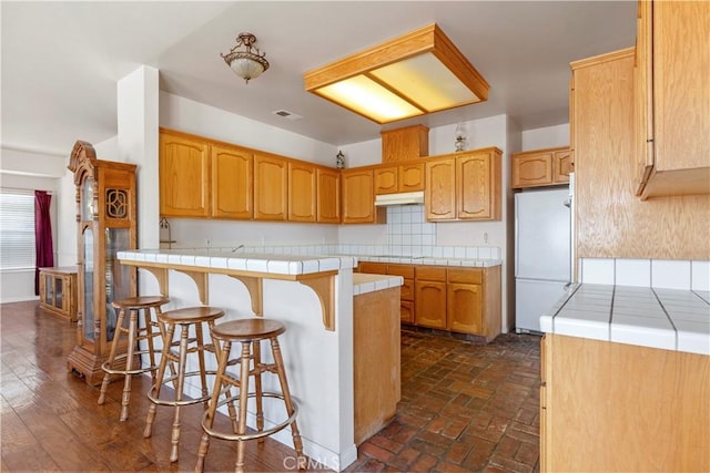 21509 Stage Dr, Tehachapi, CA 93561 - kitchen with freestanding refrigerator, tile counters, a peninsula, a kitchen bar, and backsplash