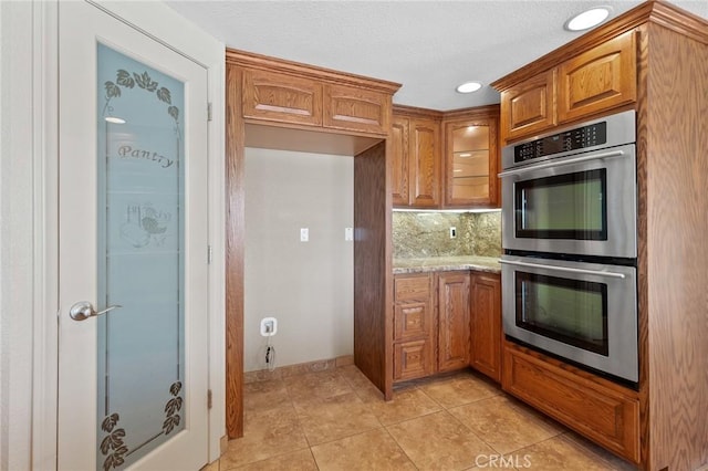 58960 Delano Trl, Yucca Valley, CA 92284 - kitchen with stainless steel double oven, brown cabinets, backsplash, light tile patterned flooring, and light stone countertops