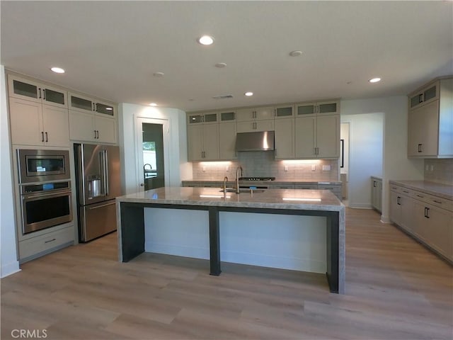 39460 Chamise St, Temecula, CA 92591 - kitchen with appliances with stainless steel finishes, under cabinet range hood, backsplash, a kitchen island with sink, and light wood-style flooring