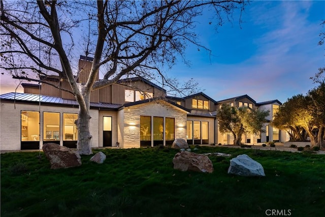 25151 Jim Bridger Rd, Hidden Hills, CA 91302 - back of property at dusk featuring a standing seam roof, a lawn, a chimney, stone siding, and a metal roof
