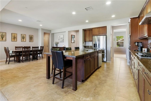 28233 Millstream Ct, Menifee, CA 92585 - kitchen featuring wall chimney exhaust hood, a kitchen bar, appliances with stainless steel finishes, an island with sink, and dark stone counters