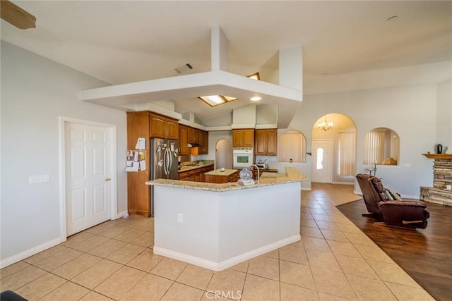 8225 Ailanthus St, Phelan, CA 92371 - kitchen featuring white oven, light tile patterned flooring, brown cabinets, arched walkways, and light stone counters