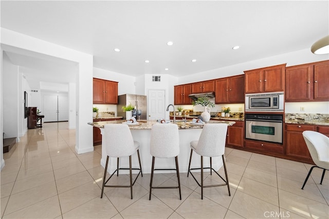 7265 Excelsior Dr, Eastvale, CA 92880 - kitchen with appliances with stainless steel finishes, under cabinet range hood, light tile patterned floors, a breakfast bar, and a center island with sink