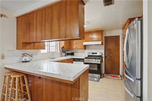 260 E Whittier Ave, Hemet, CA 92543 - kitchen featuring stainless steel appliances, under cabinet range hood, brown cabinetry, a peninsula, and tasteful backsplash
