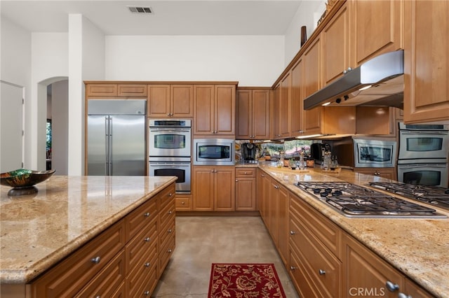 56102 Baltusrol Ln, Quinta, CA 92253 - kitchen with built in appliances, arched walkways, under cabinet range hood, light stone countertops, and brown cabinetry